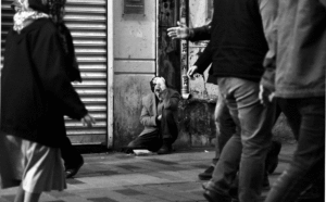 Homeless Man sitting on a dirty street, people walking by ignoring him.