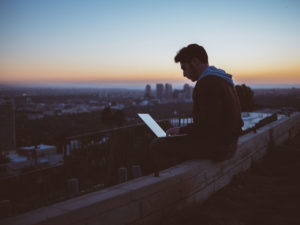 A Man Sitting on a rooftop with his laptop open, sun setting in the background.