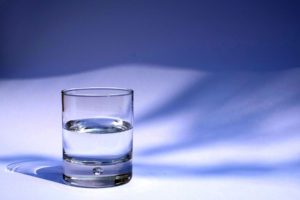 Picture of small glass of water, sitting on a white table with a dark blue wavy background.