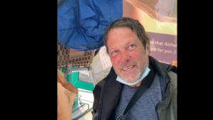 Picture of William Menn during our interview. He is seated, at a bus stop, surrounded by his belongings.