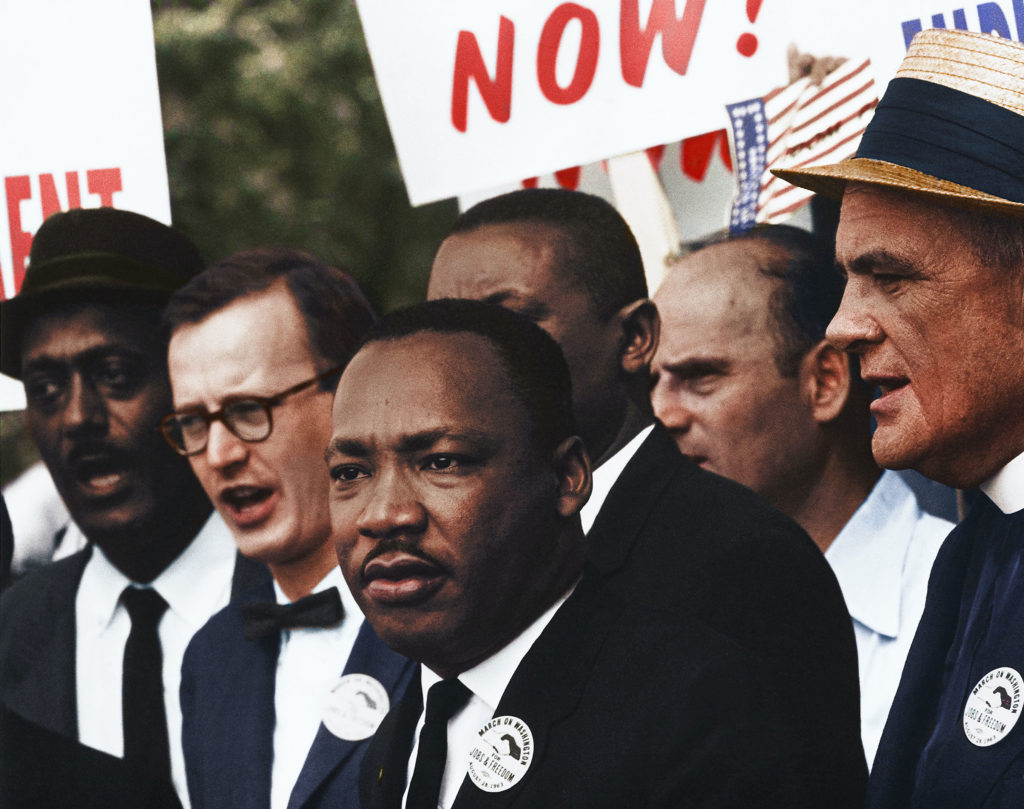 Dr. Martin Luther King Jr. speaking with a groups of men behind him. Others are holding up signs, but the only word that is visable is Now.