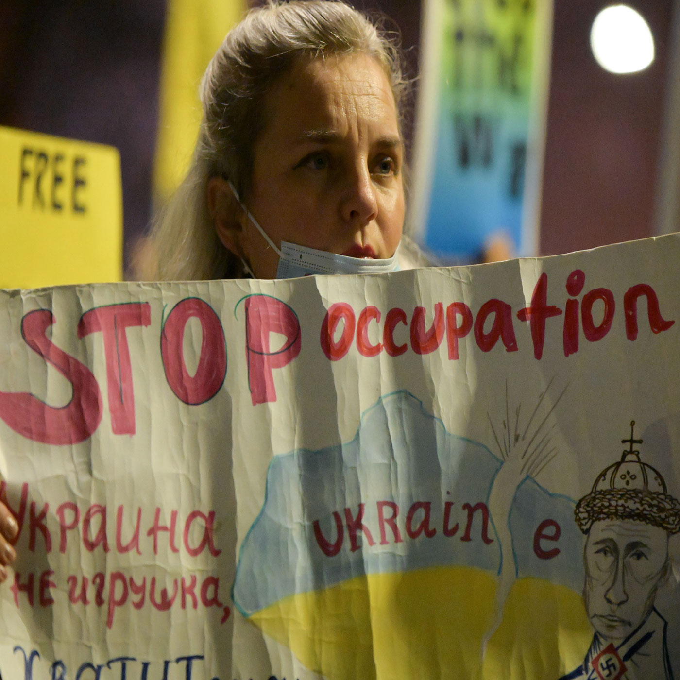 Photo is a blond woman with a mask down near her mouth, she is holding a sign that says "Stop Occupation" in red letters.